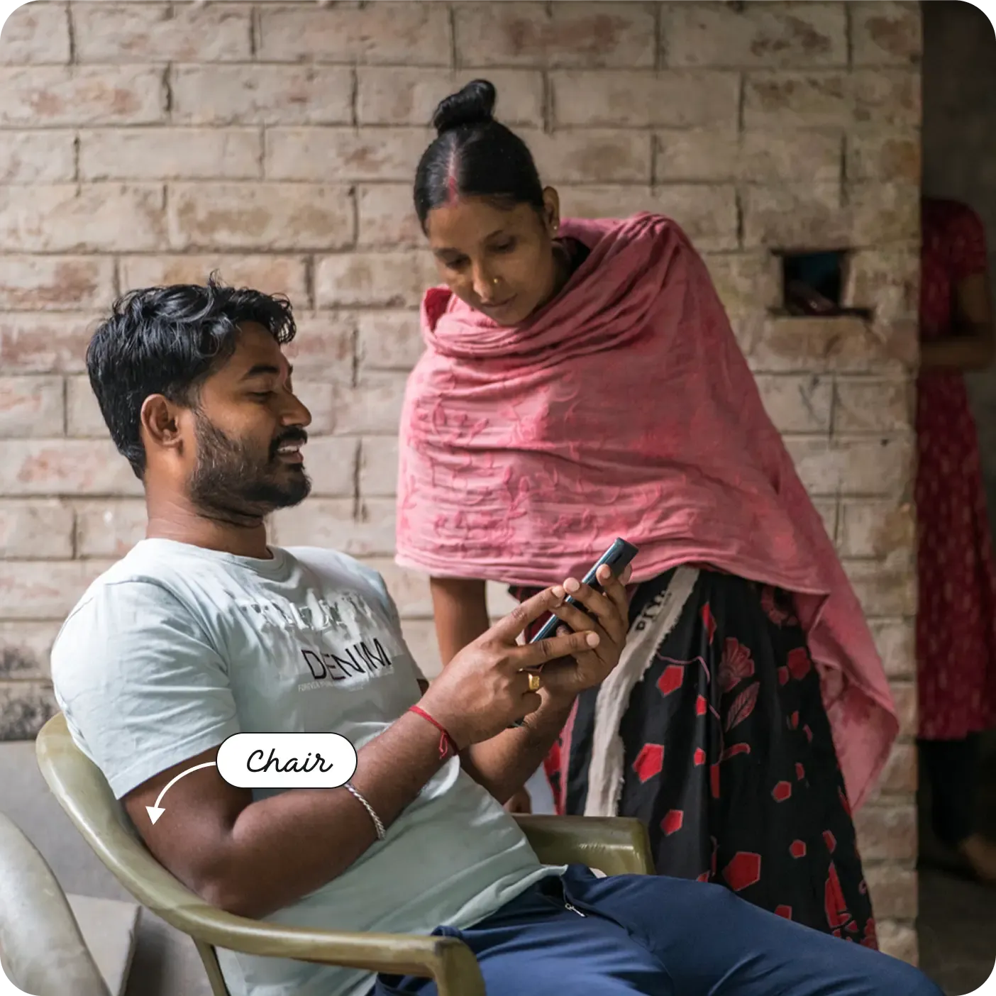 A photograph taken in front of a brick wall captures a seated Indian man looking down intently at a mobile phone. He is in a light grey chair. Standing beside and behind him, a woman wearing a pink shawl looks over his shoulder at the phone screen. A label and arrow point to the man's seat as a "Chair".