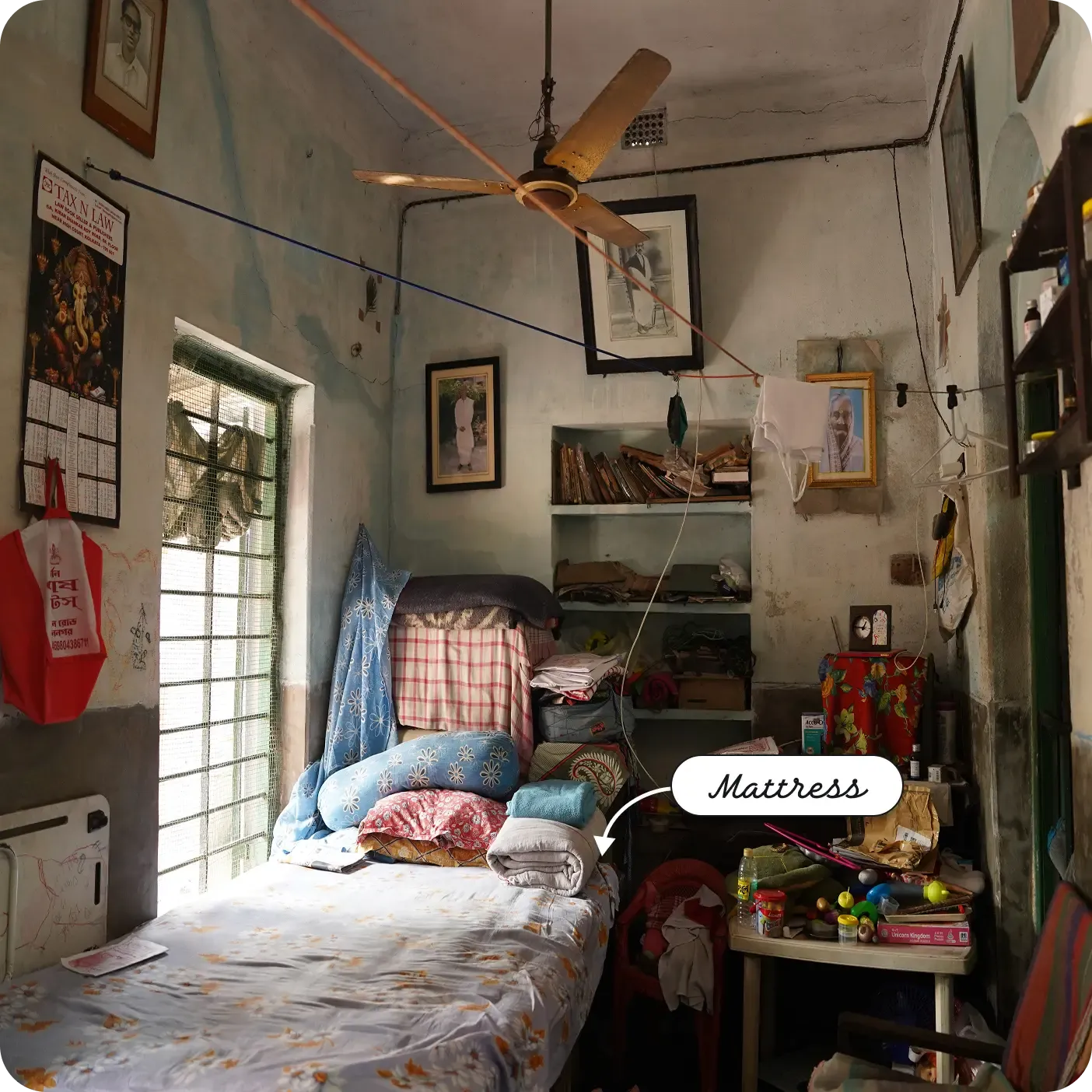 A small, lived-in room with a single bed and mattress by a window, shelves stacked with books and belongings, a ceiling fan overhead, and personal items and framed photos on the walls. A label and arrow point to the bed as a "Mattress".