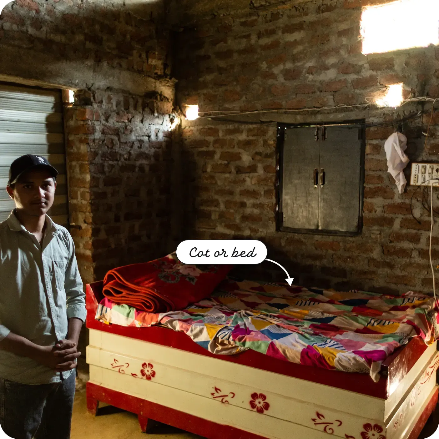 A young man stands beside a neatly made bed in a small brick room, lit by sunlight filtering through gaps in the walls. The bed is neatly made with a colorful patterned bedsheet and folded blankets, while exposed wiring and a small wall cabinet are visible on the rough brick walls. A label and arrow point to the bed as a "Cot or bed."