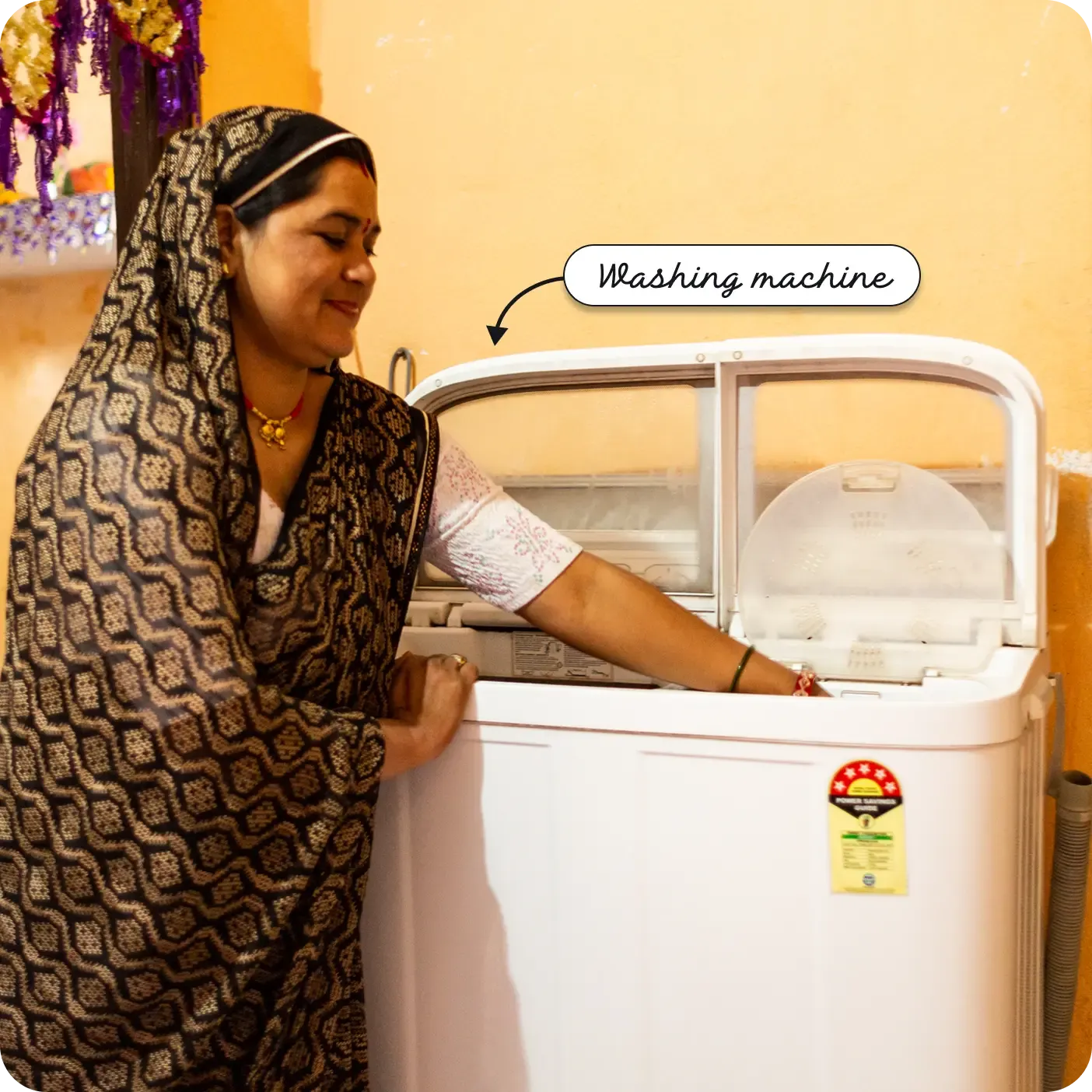 A medium close-up photograph captures an Indian woman wearing a black patterned saree and traditional jewelry, smiling as she uses a twin-tub washing machine. Her right hand is inside one of the open tubs. A yellow star-rating label is on the front of the white machine. An arrow and label point to it as a "Washing machine".