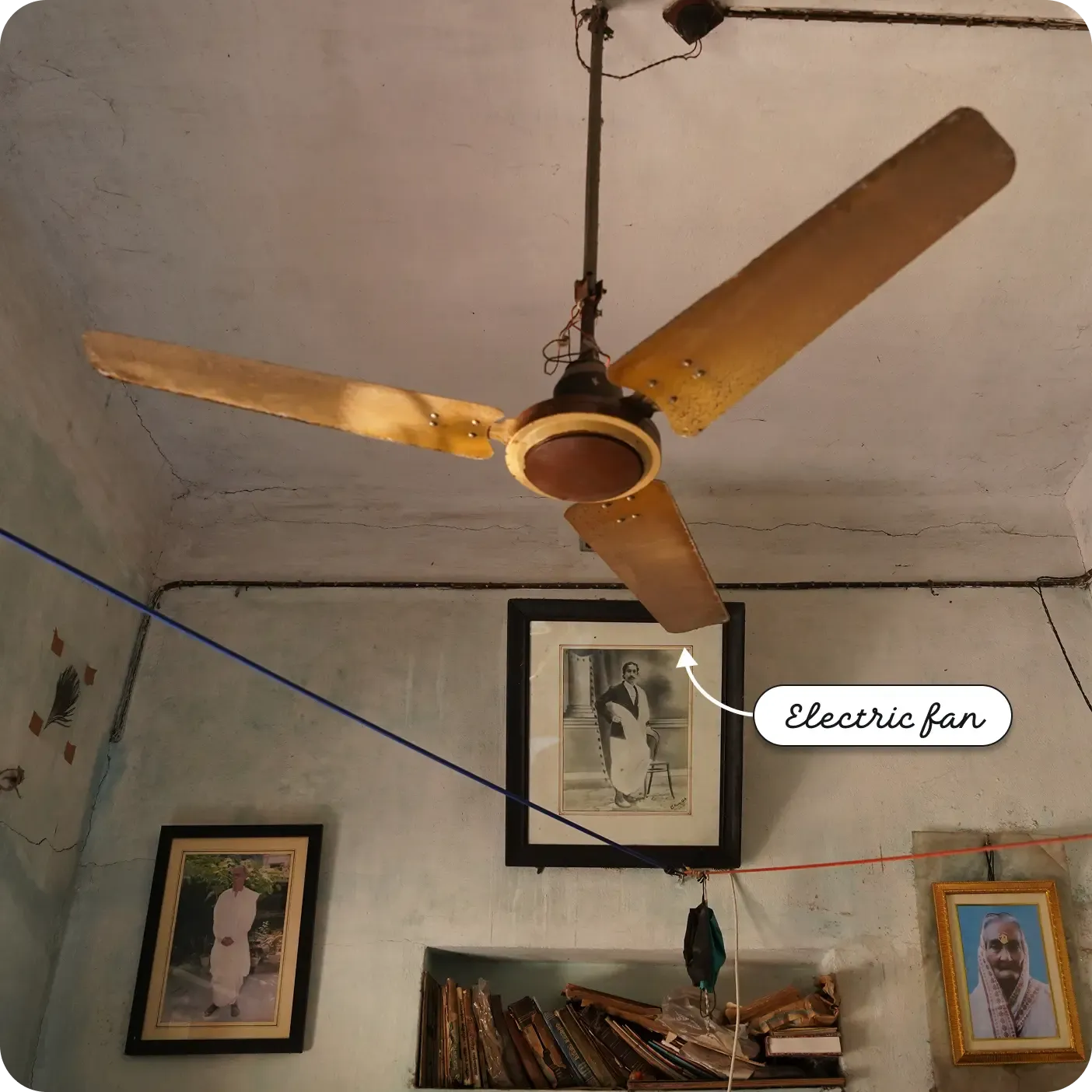 An upward-angled indoor photo focused on a three-bladed electric ceiling fan hanging from a painted ceiling. Red and blue electrical wires run across the ceiling. Below, several framed portraits and a shelf of books line the wall. An arrow and label point to the central fan unit as an "Electric fan".