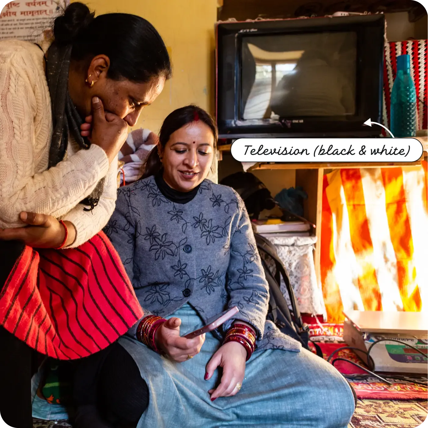 An indoor photograph showing two Indian women looking together at a smartphone. One woman is seated on the floor holding the phone and smiling, while the other stands leaning over her shoulder. Behind them on a shelf sits an older, box-style television set. A label with an arrow points to the set, reading "Television (black & white)".