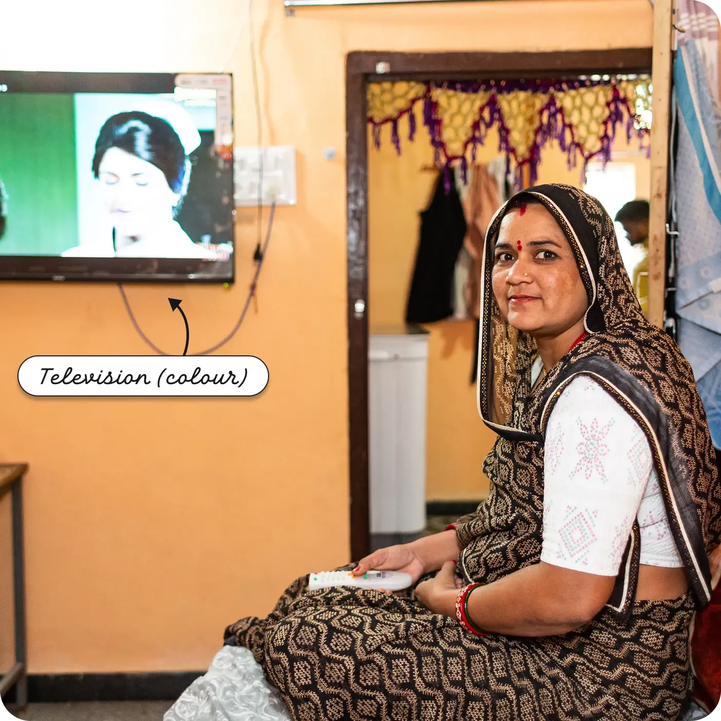 An indoor photograph of an Indian woman sitting and smiling at the camera. She wears a black and gold patterned saree and holds a white remote control in her lap. On the orange wall behind her is a mounted flat-screen TV displaying a color broadcast of a woman's face. A label and arrow point to the screen, reading "Television (colour)".