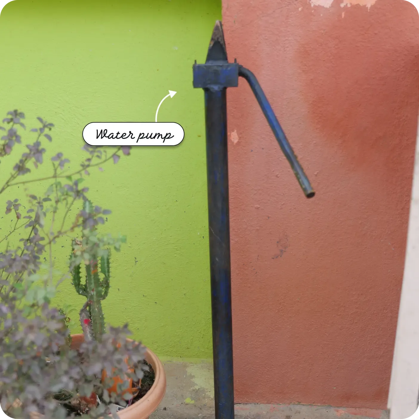 An outdoor close-up shot of a tall, blue metal manual hand water pump. It stands vertically against a background of two painted walls, one bright green and the other terracotta. A potted plant is visible in the lower left foreground. A label and arrow point to the top mechanism of the pump, reading "Water pump".