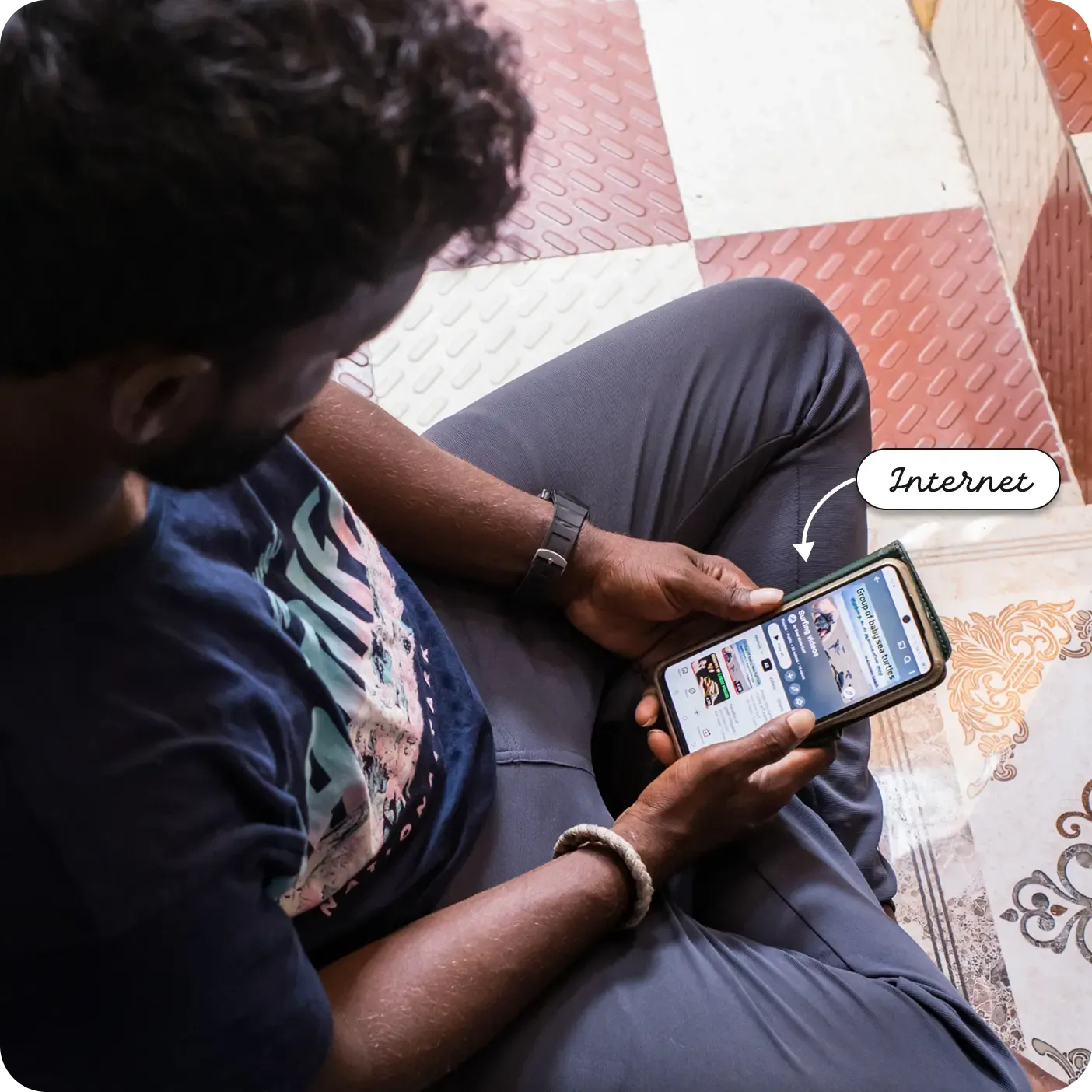 An overhead, angled photograph of a man sitting cross-legged on a patterned tile floor. He is holding a smartphone horizontally and looking at the screen, which displays an online video streaming application with thumbnails of various videos. A label and arrow point to the smartphone screen, reading "Internet".