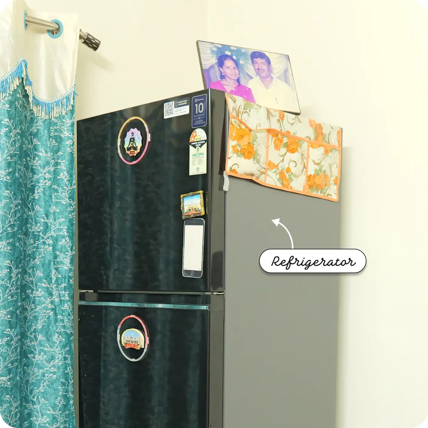 An indoor photograph showing a dark-colored, double-door refrigerator standing against a light wall next to a teal patterned curtain. The refrigerator is decorated with a few magnets and has a smartphone attached to its upper door. A floral fabric organizer hangs over the top right side, supporting a framed portrait of a couple. A label with an arrow points to the appliance, reading "Refrigerator".