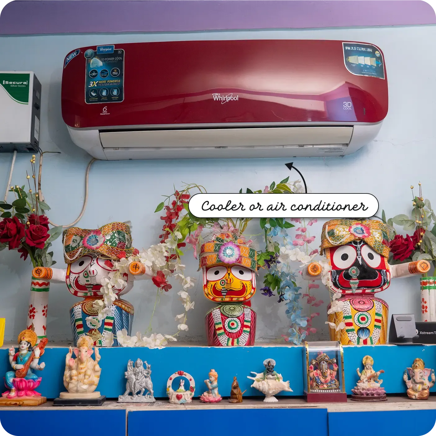 An indoor photograph of a red Whirlpool air conditioning unit mounted high on a light blue wall. Below the AC unit, a blue shelf displays three large, brightly painted wooden deities adorned with artificial flowers. A lower shelf holds several smaller religious figurines. A label and arrow point to the red unit, reading "Cooler or air conditioner".