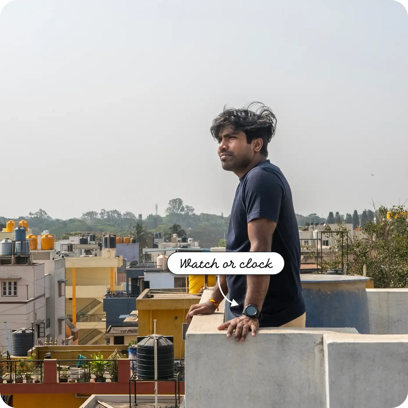 An outdoor photograph showing a man standing on a rooftop terrace, leaning his hands on a low concrete wall and looking out into the distance. He is wearing a dark blue t-shirt and has a black smartwatch on his left wrist. The background features a cityscape with buildings, black water tanks, and trees. A label and arrow point to the device on his wrist, reading "Watch or clock".