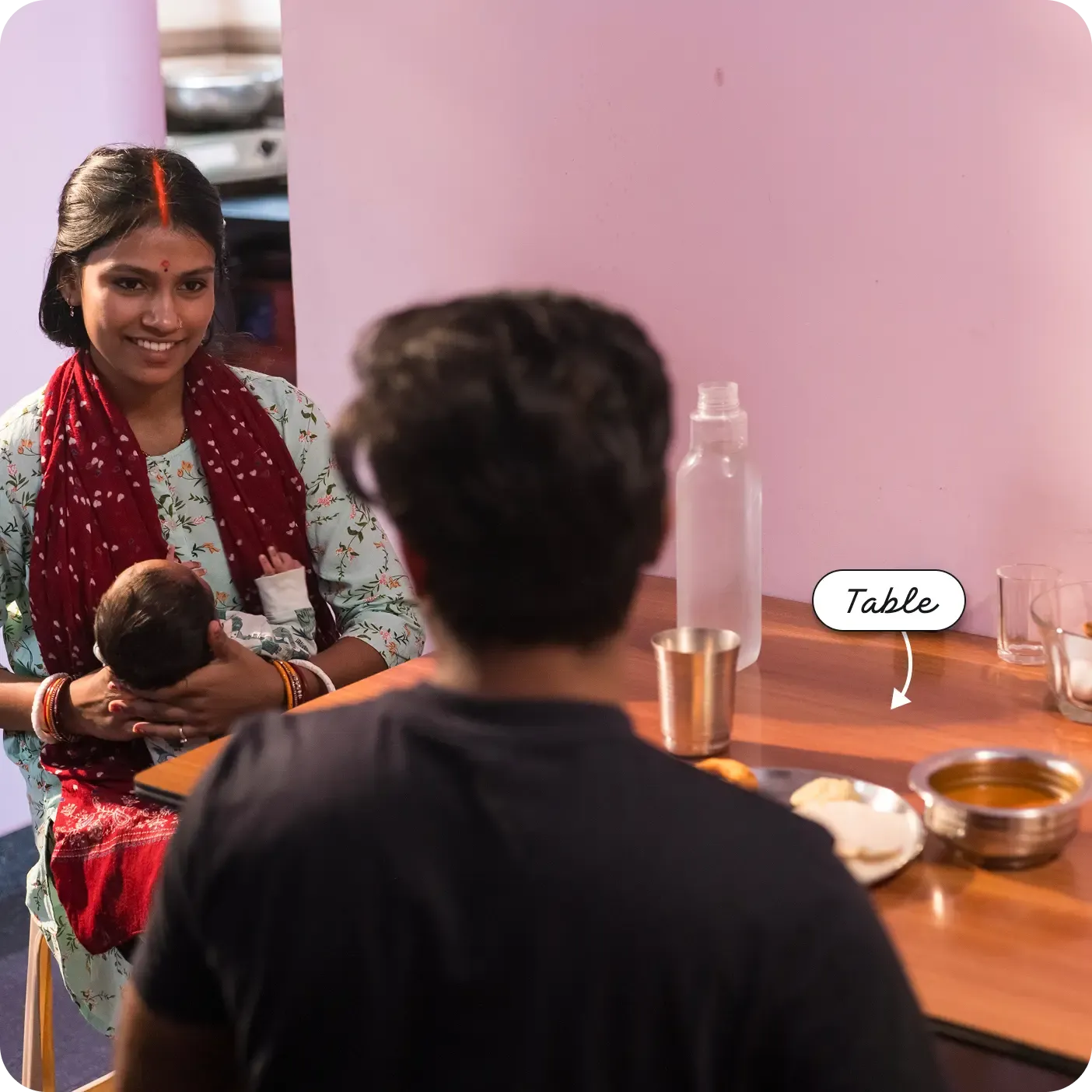 An indoor photograph capturing a woman and a man sitting across from each other at a shiny wooden dining table. The woman, facing the camera and smiling, is holding a small baby. The man is seen from behind. The table is set with a plate of food, a steel cup, a small steel bowl, and a clear water bottle. A label and arrow point to the wooden surface, reading "Table".