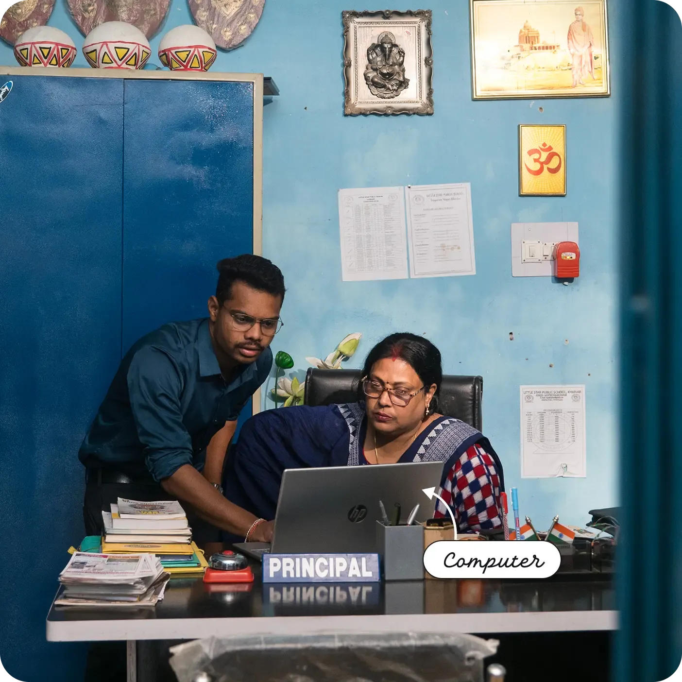 An indoor photograph of an office setting featuring a woman seated at a desk and a young man standing beside her, both looking intently at a silver HP laptop screen. The desk is cluttered with stacks of books, a bell, and a nameplate that reads "PRINCIPAL". The room has blue walls decorated with framed pictures, an Om symbol, and notices, alongside a tall blue metal cabinet. A label and arrow point to the laptop, reading "Computer".