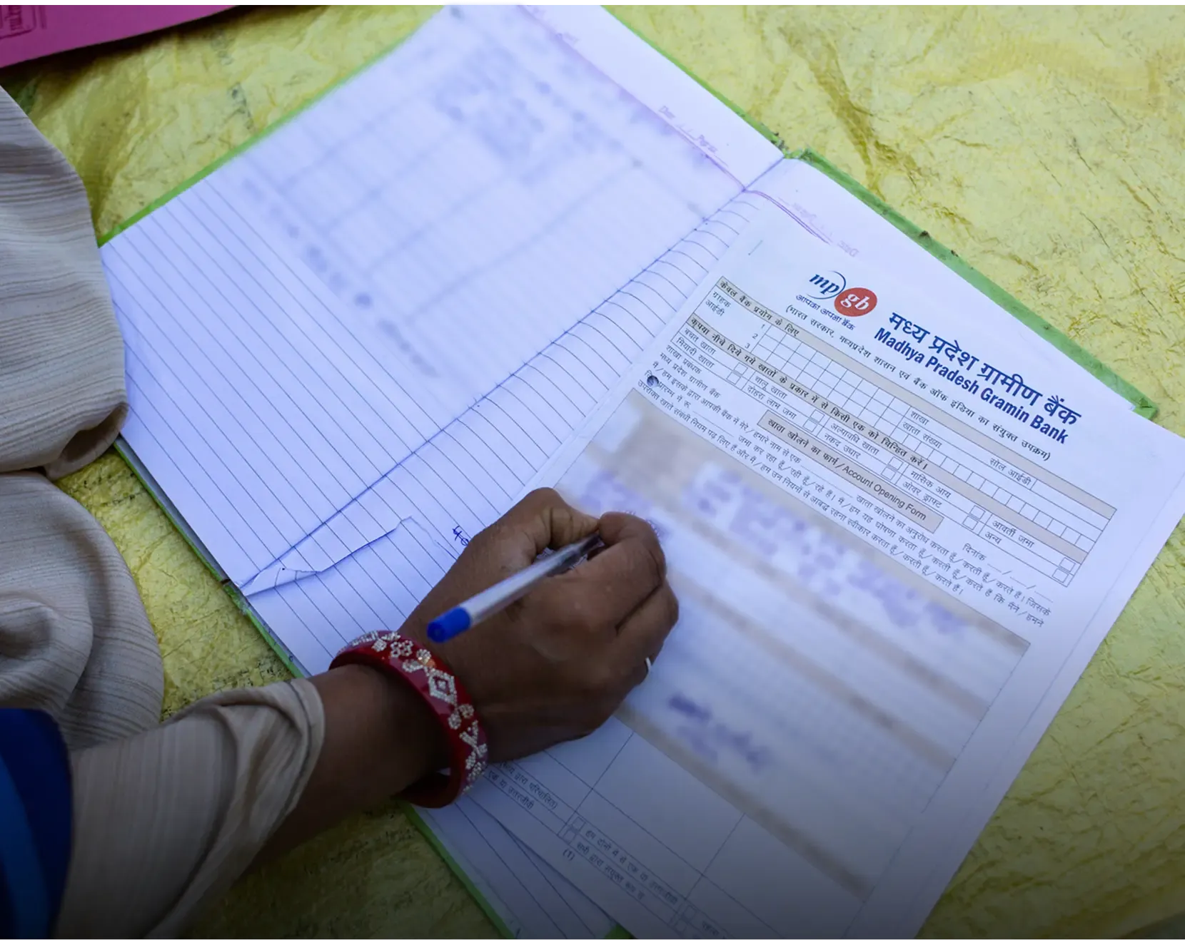 A woman's hand, adorned with red bangles, writing on a Madhya Pradesh Gramin Bank account opening form. A lined notebook is visible alongside the bank document on a yellow surface.
