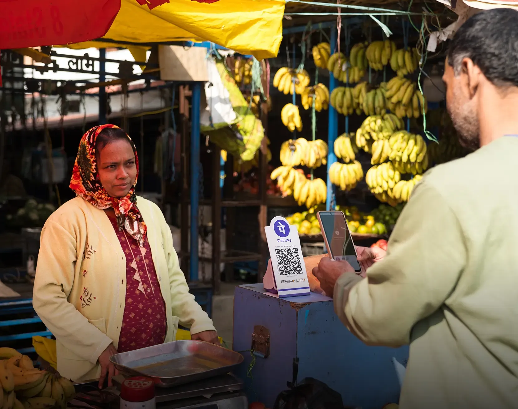 A female vendor at an outdoor fruit market stall watches as a male customer scans a PhonePe BHIM UPI QR code with his smartphone to make a digital payment. Bunches of bananas hang in the background.