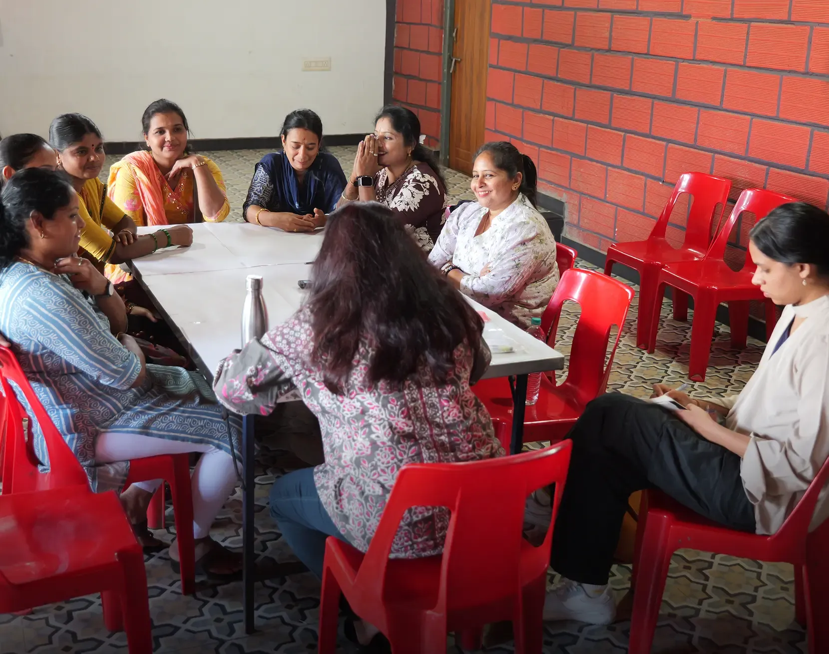 A group of approximately nine women seated around a rectangular table in a room with red brick walls and red plastic chairs, engaged in a group discussion.