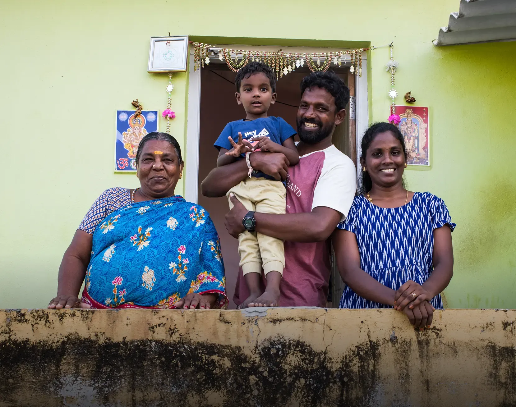 A smiling family of four — an older woman in a blue saree, a man holding a young boy—his son, and a younger woman—his wife, posing together on the porch of a light green home decorated with religious imagery and decorative garlands.