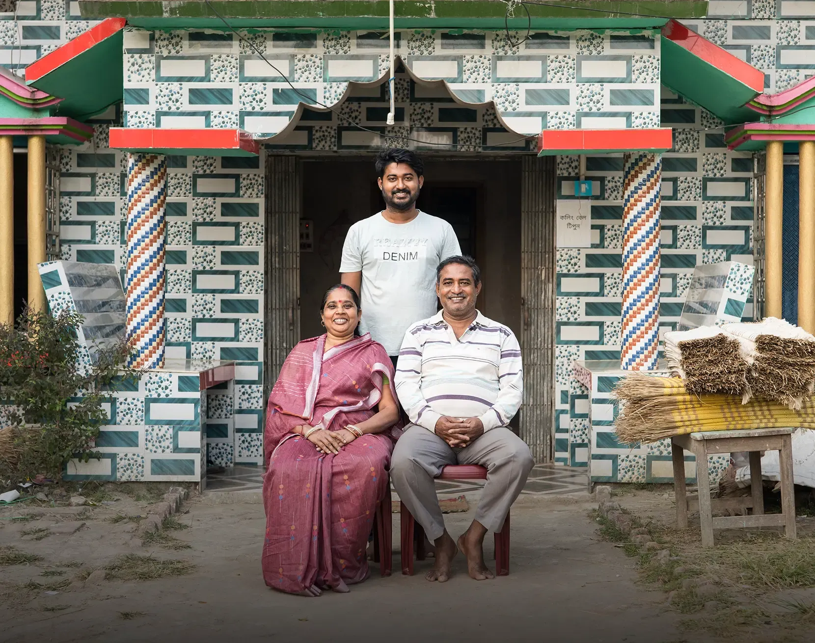 A family of three — a woman in a pink saree, a seated man in a striped shirt, and a young man standing behind them — smiling in front of a vibrantly decorated home with colorful tile-patterned walls.