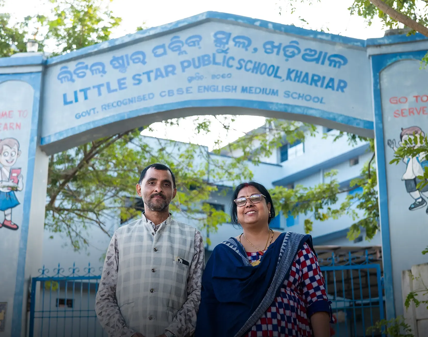 A man and a woman standing together in front of the entrance gate of Little Star Public School in Khariar, a government-recognised CBSE English medium school, with trees visible in the background.